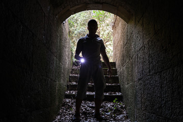 Young man with a flashlight enters stone tunnel - obrazy, fototapety, plakaty