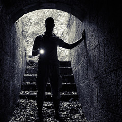 Young man with a flashlight enters the stone tunnel - obrazy, fototapety, plakaty