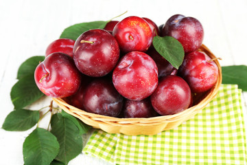 Ripe plums in wicker bowl on wooden table with napkin, closeup