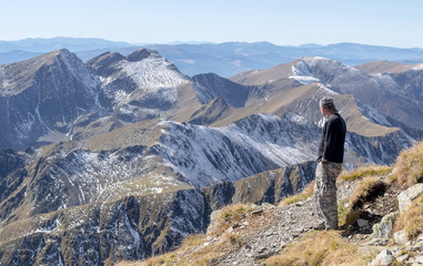 Lonely man stands on top of a mountain and looking into the distance on a background of mountain range. Focus on the first plan, on the man. Mountains on the background is blurry.