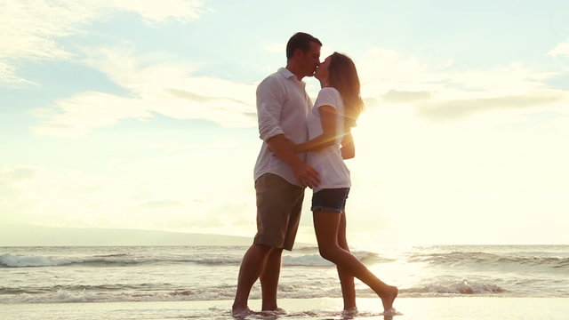 Honeymoon passionate couple kissing on the beach. Beautiful Sunset Flares. Romantic Newlywed happy young couple enjoying ocean sunset during travel holidays vacation getaway.  Shot with Steadicam.