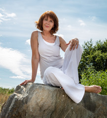 Fototapeta na wymiar senior zen - smiling, beautifully aging woman sitting on a stone for outdoors yoga session wearing white seeking serenity and wellness in a park,summer daylight