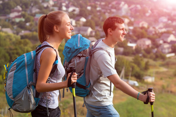 Excited Travelers Young Man and Woman Traveling Outdoor Expressing Fun and Pleasure with Backpacks Walking Poles Sticks and Casual Sporty Style Clothing Suburban Park Landscape Shining Sun Background