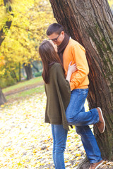 Fototapeta premium Young man kissing a girl in a park on a sunny autumn day while leaning on a tree.