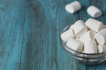 Fototapeta na wymiar White soft marshmallows in glass bowl on blue wooden background, selective focus