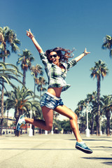 Beautiful young crazy happy girl is jumping in palm trees in summer in Barcelona wearing sunglasses blue jeans shirts - 90826112