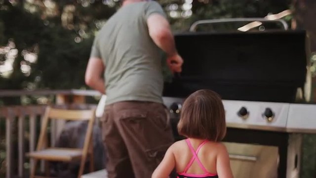 A little girl watches her dad clean a barbecue

