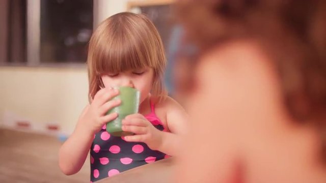 A little girl drinking from a cup when her mom brings her a hot dog and chips