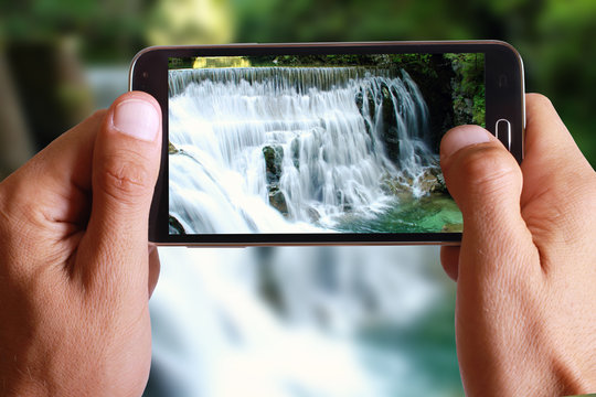 Male Hand Taking Photo Of A Green Rock Mountain With A Waterfall Landscape With Cell, Mobile Phone.