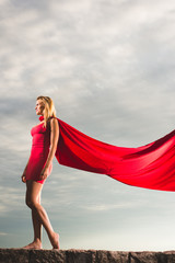 Blonde woman in red dress and red mantle posing outdoor as a superhero against blue sky