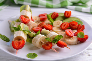 Fresh fruits on skewers in plate on wooden table, closeup