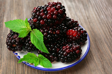 Ripe blackberries with green leaves in saucer on wooden table, closeup