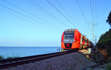 Электричка движется по железнодорожному пути вдоль берега моря в освещении утреннего солнца
