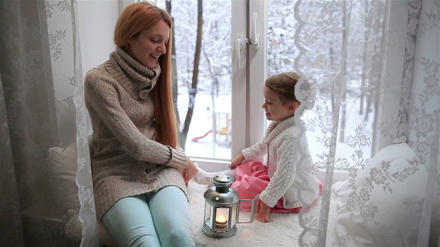 Young mother and her adorable daughter lit Christmas lantern on a window sill. Dolly shot.
