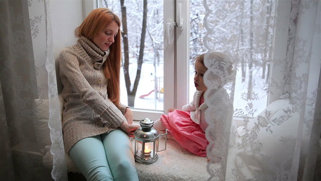 Young mother and her adorable daughter lit Christmas lantern on a window sill. Dolly shot.