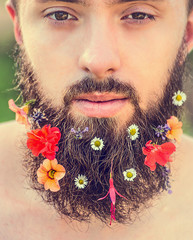 man's face with a beard with flowers in his beard on natural background, close up