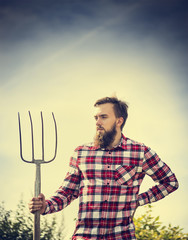 Portrait of young bearded farmer in red checkered shirt with old pitchfork on sky  nature backgrund, toned
