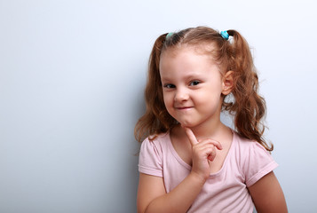 Cunning thinking small kid girl with finger near face on blue ba