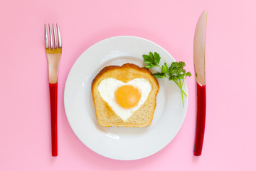 Heart shaped egg in toast. White plate, red handles, pink background. Top view.