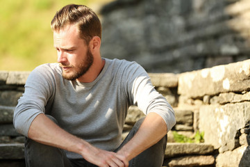 Man in his 20s wearing a grey shirt and jeans, sitting down outside on a set of steps on a sunny...