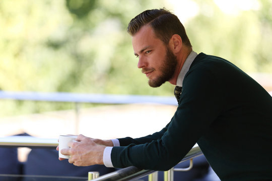 A pensive man wearing a pullover and holding a coffe mug, standing on a patio on a sunny summer day.