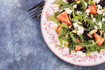 Diet or vegetarian concept. Fresh salad with arugula, cherry, strawberry, feta cheese on blue rustic background. Top view. Selective focus