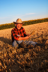 Naklejka na ściany i meble Senior farmer sitting in a wheat field after harvest and examining crop