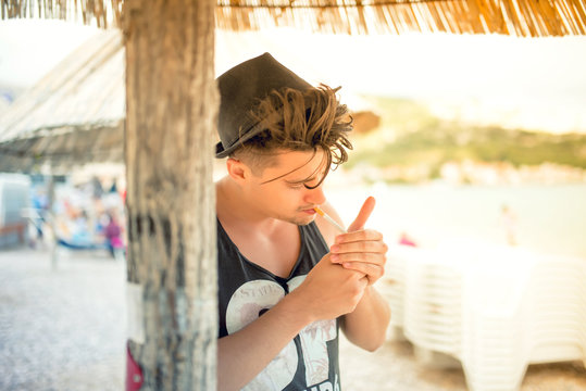 portrait of male hipster lighting a cigarette on the beach, wearing hat and tank top. concept of happy vacation