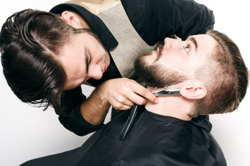 Obraz premium Young hairdresser shaving brutal bearded customer with razor