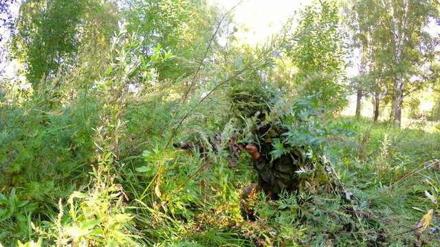 sniper with a gun camouflaged in the grass in forest searching victim