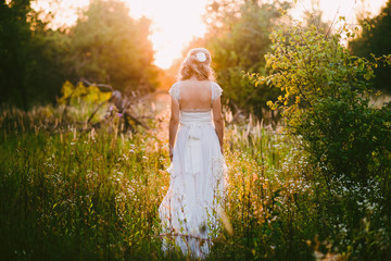 Stylish beautiful blonde bride standing in her wedding dress back to nature in the sunset light,...