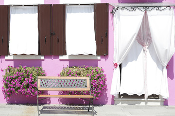 Pink painted wall with two windows with wooden shutters and a door covered with a curtain on the windowsill, a potted with flowers, at the island of Burano, Venice, Italy