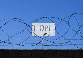 Flüchtling hinter Stacheldraht hält Schild mit Aufschrift HOPE hoch