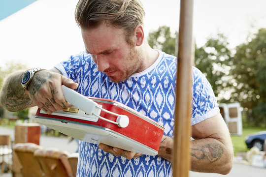 A man in a blue shirt with tattooed forearms, looking at a red vintage radio at a flea market.