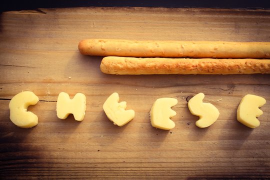 The word cheese written in white cheese on a wooden surface with bread sticks