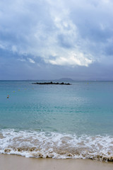 Die Sand aufwirbelnde Welle des blauen unter einer Wolkendecke liegenden Meeres am Sandstrand...