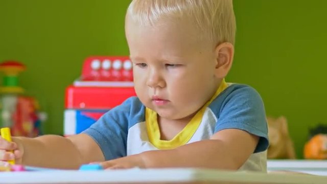 Toddler boy practice to paint with crayons on paper sitting at the table in child room