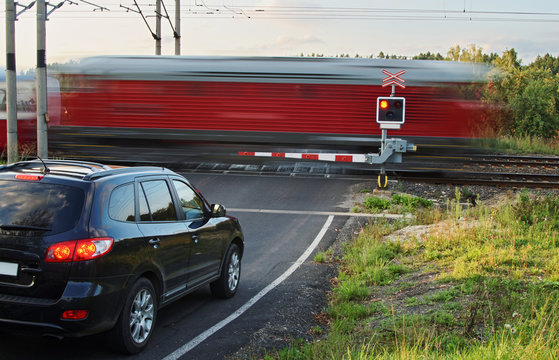 Fototapeta Speeding motion blur red train passing through a railway crossing with gates. Black car standing in front of the railway barriers on an asphalt road.
