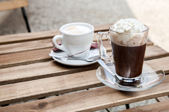 The cup of hot cocoa with whipped cream and cup of capuccino are on the table in outdoor cafe