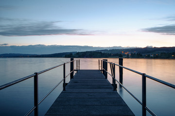 Ein Bootssteg führt in den See. Die Nacht weicht dem Morgenrot.
