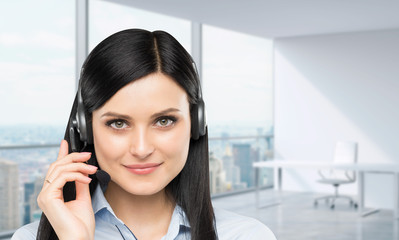 Front view of the smiling brunette support phone operator with headset. Panoramic office workplace...