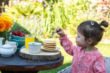 Toddler girl having breakfast in the garden: pancakes with yoghurt and berries on a rustic table