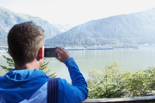 White Male Tourist Taking Picture of Cruise Ships next to mountain in Alaskan River with Cell Phone