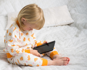 Маленькая девочка сидит на постели в пижамке и смотрит электронную книгу. Электронная книга.