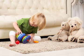 Ребенок сидит на полу, на коврике и собирает игрушечную пирамидку из разноцветных колец. В комнате красиво и уютно. Рядом с малышом сидят игрушечные белые медведи