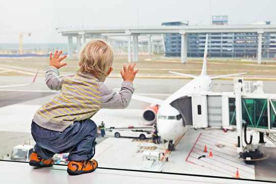 Little Baby Boy Waiting Boarding To Flight In Airport Transit Hall And Looking Through The Window At Airplane Near Departure Gate. Active Lifestyle, Travel By Air With Child On Family Summer Vacation