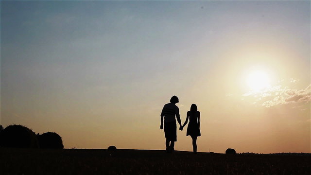 Young slim and playful silhouette couple standing and walking in a field full of haystacks at sunset