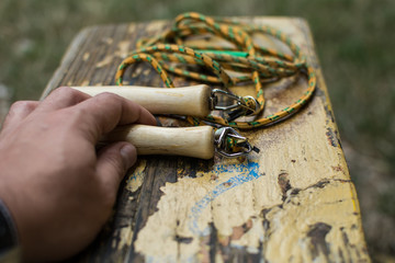 Children jump rope on a park bench. In the focus wooden handle and metal noose. Man hand is holding one handle.