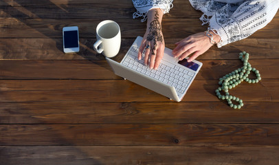 Female Hands Working on Laptop Natural Wooden Desk with White Computer Large Mug Telephone and Rosary Woman Typing on Keyboard with Stylish Tattoo on Wrist Top View