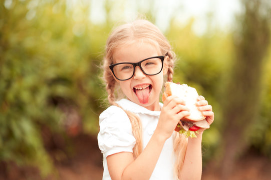 Funny kid girl eating sandwich outdoors. Having fun. Looking at camera. Posing over nature background. Healthy food. Childhood. 